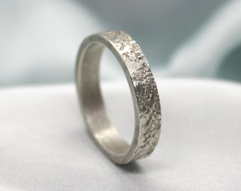 May include: A silver ring with a textured surface. The ring has a band with a rough, uneven texture, giving it a unique, organic appearance. The ring is set against a soft, white background.