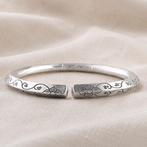 May include: A silver-toned open cuff bracelet featuring a polished finish and engraved with intricate floral patterns and swirling vine motifs along the outer surface. The bracelet has a rounded profile and a gap at the front for easy wear.