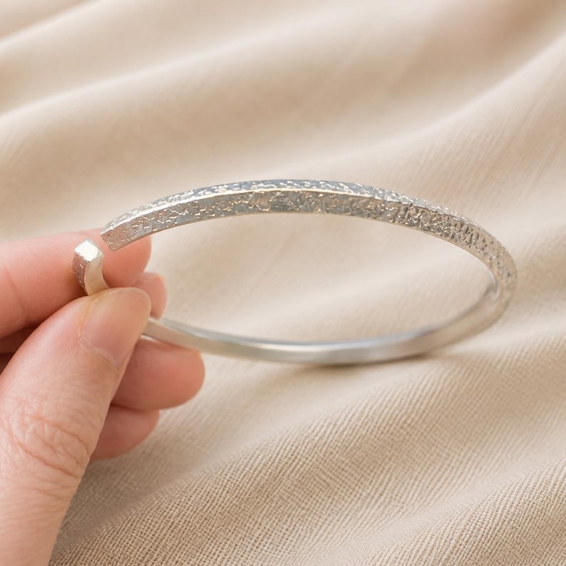 Puede incluir: Una pulsera tipo brazalete de tono plateado con una superficie exterior texturizada y rugosa, y una banda interior lisa y pulida. La pulsera es abierta y presenta un acabado met&aacute;lico con un contraste marcado entre el exterior y el interior.