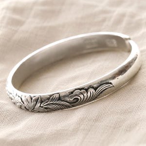 Puede incluir: Una pulsera r&iacute;gida de metal en tono plateado que presenta un patr&oacute;n continuo de flores y hojas en relieve alrededor del exterior. La superficie tiene un acabado pulido con &aacute;reas hundidas oscurecidas que resaltan los detalles bot&aacute;nicos.