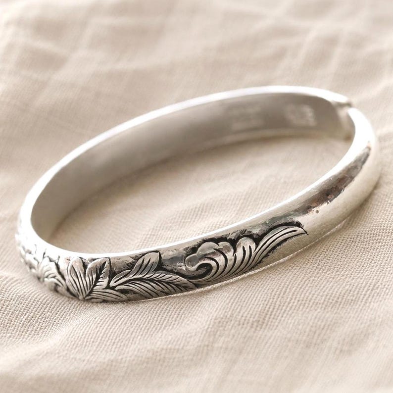 Puede incluir: Una pulsera r&iacute;gida de metal en tono plateado que presenta un patr&oacute;n continuo de flores y hojas en relieve alrededor del exterior. La superficie tiene un acabado pulido con &aacute;reas hundidas oscurecidas que resaltan los detalles bot&aacute;nicos.