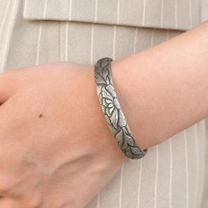 Puede incluir: Una pulsera r&iacute;gida de metal en tono plateado con un patr&oacute;n floral en relieve que muestra p&eacute;talos y hojas detallados. La pulsera tiene un acabado pulido con &aacute;reas hundidas oscurecidas que resaltan el intrincado dise&ntilde;o bot&aacute;nico.
