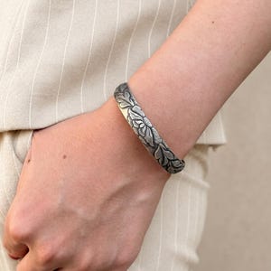 Puede incluir: Una pulsera r&iacute;gida de plata con un intrincado patr&oacute;n floral y de hojas en relieve, con detalles oxidados en las zonas hundidas. La superficie met&aacute;lica tiene un acabado pulido en las partes elevadas, resaltando el dise&ntilde;o bot&aacute;nico.