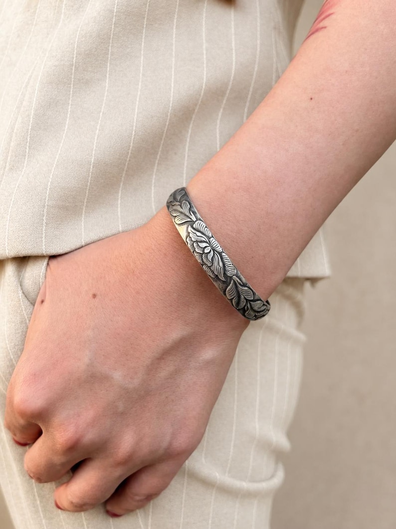 Puede incluir: Una pulsera r&iacute;gida de plata con un intrincado patr&oacute;n floral y de hojas en relieve, con detalles oxidados en las zonas hundidas. La superficie met&aacute;lica tiene un acabado pulido en las partes elevadas, resaltando el dise&ntilde;o bot&aacute;nico.