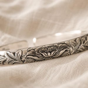Puede incluir: Una pulsera tipo brazalete de plata con un patr&oacute;n floral detallado en relieve en su superficie exterior. El metal tiene un acabado pulido con &aacute;reas hundidas oscurecidas que resaltan los intrincados dise&ntilde;os de p&eacute;talos y hojas en la banda.