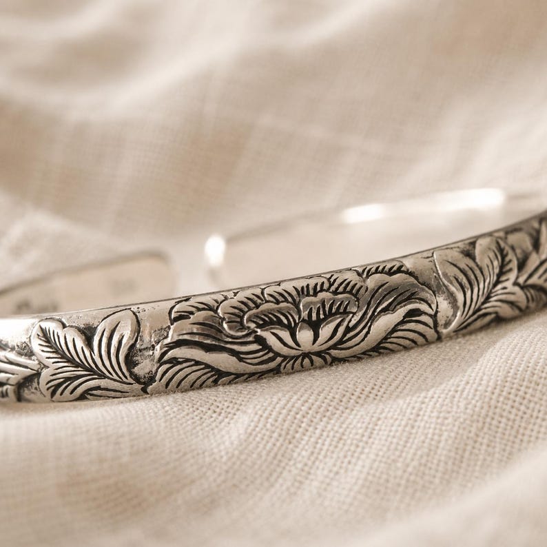 Puede incluir: Una pulsera tipo brazalete de plata con un patr&oacute;n floral detallado en relieve en su superficie exterior. El metal tiene un acabado pulido con &aacute;reas hundidas oscurecidas que resaltan los intrincados dise&ntilde;os de p&eacute;talos y hojas en la banda.