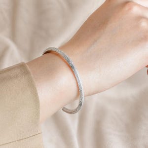 Puede incluir: Una pulsera tipo brazalete de tono plateado con una superficie texturizada y granulada. La pulsera presenta un dise&ntilde;o delgado y abierto que rodea la mu&ntilde;eca, mostrando un acabado met&aacute;lico consistente con una apariencia fina y arenosa en el exterior.