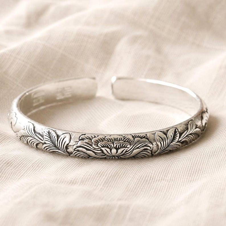 Puede incluir: Una pulsera abierta de tono plateado que presenta un patr&oacute;n floral en relieve detallado en la superficie exterior. El metal tiene un acabado pulido con &aacute;reas hundidas oscurecidas que resaltan los intrincados dise&ntilde;os de p&eacute;talos y hojas.