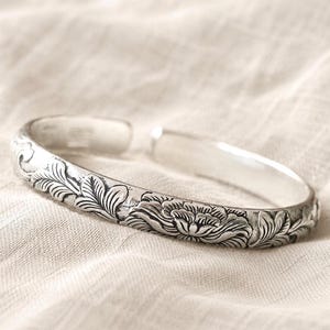Puede incluir: Una pulsera abierta de tono plateado con un patr&oacute;n detallado en relieve de flores en flor y follaje. La superficie met&aacute;lica tiene un acabado pulido con &aacute;reas hundidas oscurecidas que resaltan los intrincados grabados florales.