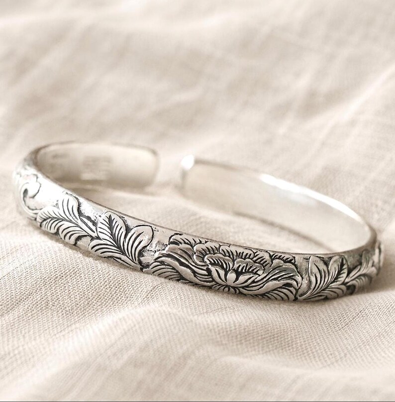 Puede incluir: Una pulsera abierta de tono plateado con un patr&oacute;n detallado en relieve de flores en flor y follaje. La superficie met&aacute;lica tiene un acabado pulido con &aacute;reas hundidas oscurecidas que resaltan los intrincados grabados florales.