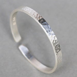 May include: A silver-colored open ring featuring a textured, hammered finish on the outer surface. The band has a slim, uniform width and a gap at one side, creating an adjustable design with a metallic sheen and reflective highlights.