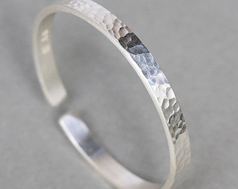 May include: A silver-colored open ring featuring a textured, hammered finish on the outer surface. The band has a slim, uniform width and a gap at one side, creating an adjustable design with a metallic sheen and reflective highlights.