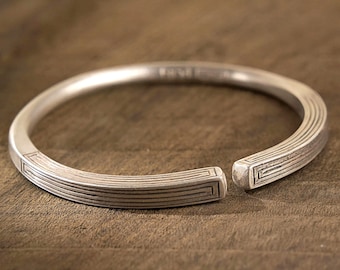 May include: A silver-colored open cuff bracelet. The bracelet has a geometric design with parallel lines and rectangular accents. The bracelet is displayed on a wooden surface.