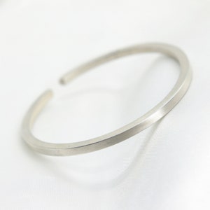 Puede incluir: Una pulsera tipo brazalete de metal en tono plateado con un acabado mate liso y un diseño circular abierto sencillo. La pulsera presenta una superficie limpia y sin adornos con un grosor constante en toda su circunferencia.