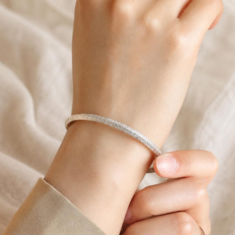 Puede incluir: Una pulsera r&iacute;gida delgada de tono plateado con un acabado de superficie texturizado y granulado. La banda de metal tiene un perfil redondeado y un ancho constante, rodeando la mu&ntilde;eca con un brillo sutil y reflectante en toda su superficie.