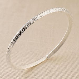 Puede incluir: Un brazalete de color plateado que presenta una superficie exterior texturizada y rugosa con un interior liso y pulido. El brazalete tiene forma circular con una peque&ntilde;a abertura en un extremo, mostrando un acabado met&aacute;lico y un dise&ntilde;o fino.