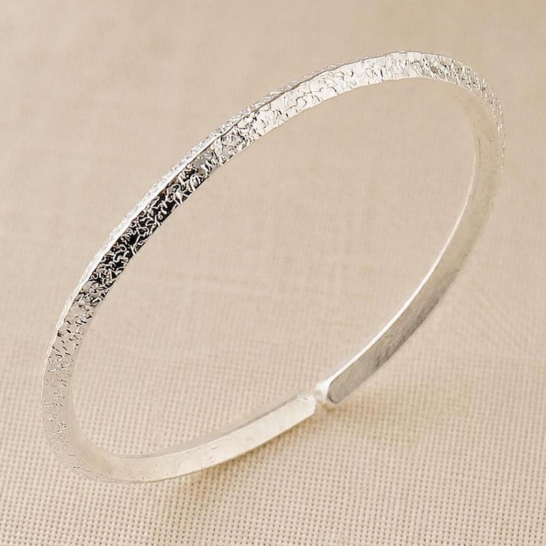 Puede incluir: Un brazalete de color plateado que presenta una superficie exterior texturizada y rugosa con un interior liso y pulido. El brazalete tiene forma circular con una peque&ntilde;a abertura en un extremo, mostrando un acabado met&aacute;lico y un dise&ntilde;o fino.