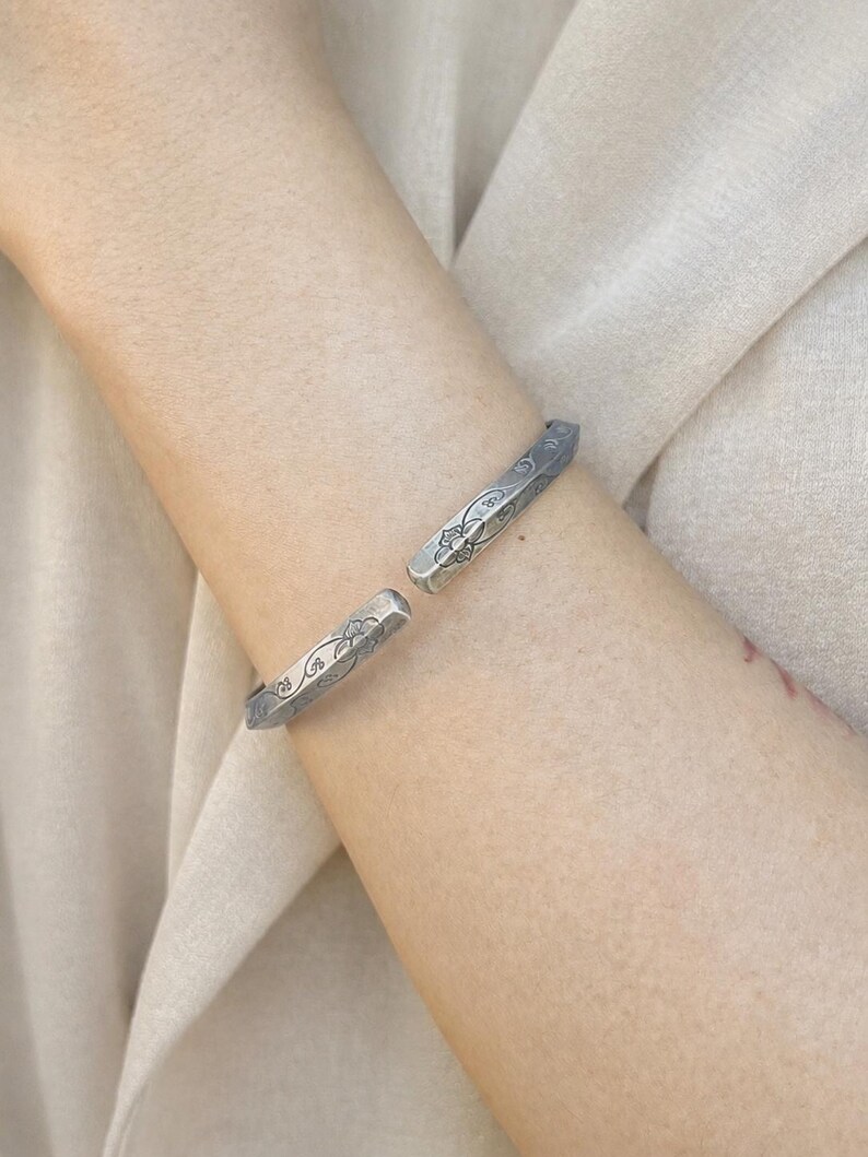 May include: A silver-toned open cuff bracelet featuring a faceted, angular profile. The surface of the metal is decorated with an engraved floral pattern consisting of delicate petals and swirling vine motifs along the outer edges of the band.