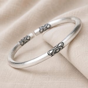 May include: A silver-toned open cuff bracelet with a brushed metallic finish. The bracelet features two distinct sections of dark, recessed decorative engravings that depict swirling cloud-like patterns near the open ends of the circular band.
