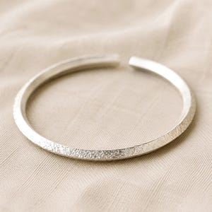 Puede incluir: Una pulsera abierta de color plateado que presenta una superficie texturizada y granulada en la banda exterior y un acabado liso y pulido en el lado interior. La pieza circular tiene una peque&ntilde;a abertura en la parte superior, formando un aro.
