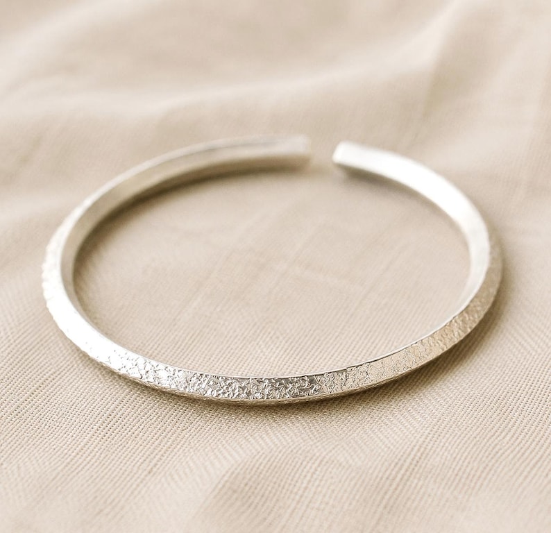 Puede incluir: Una pulsera abierta de color plateado que presenta una superficie texturizada y granulada en la banda exterior y un acabado liso y pulido en el lado interior. La pieza circular tiene una peque&ntilde;a abertura en la parte superior, formando un aro.