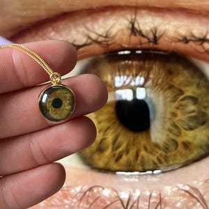 Könnte beinhalten: Eine goldfarbene Anhängerkette mit einem runden Anhänger, der ein realistisches, detailliertes haselnussbraunes menschliches Auge zeigt. Der Anhänger hängt an einer feinen goldfarbenen Kette und präsentiert ein hochauflösendes Irismuster.