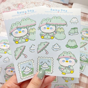 May include: A set of 10 cute penguin and frog themed stickers with a rainy day theme. The stickers are on a blue background with white clouds and raindrops. The stickers feature a penguin wearing a raincoat and holding an umbrella, a frog wearing a raincoat, a paper boat, and a pair of rain boots. The stickers are handmade with love by Gellyroise.