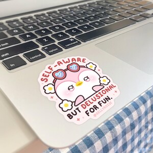 Puede incluir: Una pegatina de ping&uuml;ino rosa con gafas de sol y una flor en la cabeza. La pegatina dice "Self-Aware But Delusional For Fun." y tiene el nombre del artista, "@GellyRose", en la parte inferior.