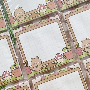 Puede incluir: Blocs de notas cuadrados con un patr&oacute;n de cuadr&iacute;cula y un borde decorativo con osos de dibujos animados, setas y plantas en macetas en tonos marr&oacute;n, rosa y verde. Los blocs de notas est&aacute;n empaquetados en pl&aacute;stico transparente.