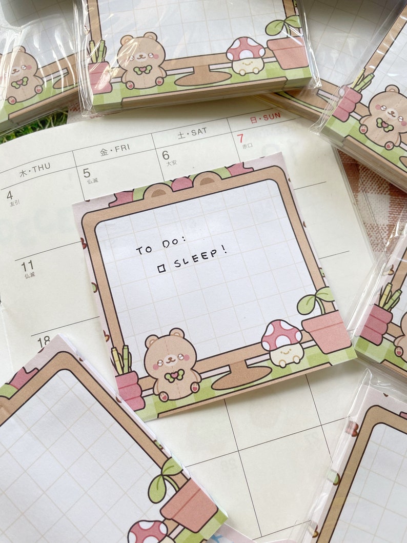 Puede incluir: Blocs de notas cuadrados con un patr&oacute;n de cuadr&iacute;cula y el texto "To Do: SLEEP!". Cada bloc de notas presenta un dise&ntilde;o de oso de dibujos animados, hongo y plantas en macetas en tonos marrones, rosas y verdes. Algunos blocs de notas est&aacute;n en envases de pl&aacute;stico transparente.