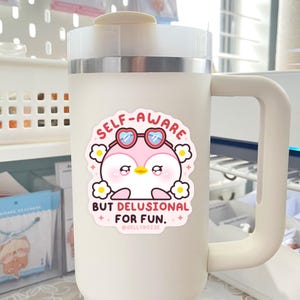 Puede incluir: Taza de viaje blanca con una pegatina de ping&uuml;ino rosa que dice "Self-Aware But Delusional For Fun." La pegatina tambi&eacute;n incluye gafas de sol en forma de coraz&oacute;n rosa y margaritas blancas.