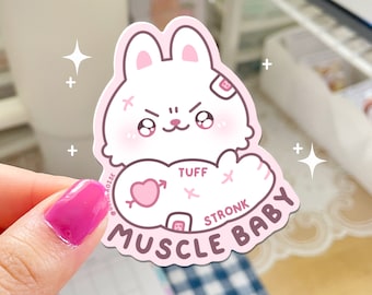 Puede incluir: Pegatina rosa y blanca con un conejito de dibujos animados con un vendaje en la cabeza. El conejito sostiene un corazón con una flecha apuntando hacia la derecha. El texto "Tuff" y "Stronk" están por encima del corazón. El texto "Muscle Baby" está debajo del corazón.