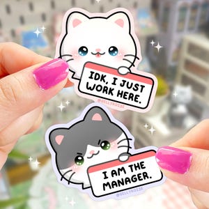 May include: Two cat stickers with name tags. The white cat sticker has a pink name tag that says "IDK, I JUST WORK HERE." The gray cat sticker has a pink name tag that says "I AM THE MANAGER."