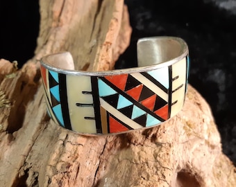 May include: A silver cuff bracelet with a geometric pattern of turquoise, red, black, and white stones.