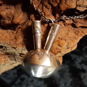 Op de afbeelding: Een zilveren hangerketting met een uniek ontwerp. De hanger heeft een afgeronde basis met geëtste patronen en twee rechtopstaande, cilindrische elementen. De ketting is ook zilverkleurig. De hanger wordt gepresenteerd tegen een getextureerde, bruine achtergrond.