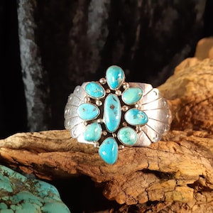 Può includere: Un bracciale a polsino in argento con pietre turchesi. Il bracciale presenta un gruppo centrale di pietre turchesi ovali e a goccia, incastonate in una fascia d'argento con un design dettagliato e strutturato. Le pietre turchesi sono di un vivace colore blu-verde.