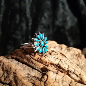 Könnte beinhalten: Ein silberner Ring mit einem floralen Design, besetzt mit türkisfarbenen Steinen. Der Ring liegt auf einem Stück verwittertem Holz vor dunklem Hintergrund.