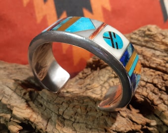 May include: A silver cuff bracelet with a geometric design inlaid with turquoise, lapis lazuli, coral, and other stones.