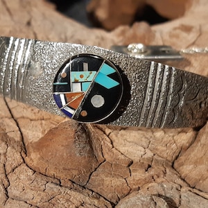 Op de afbeelding: Een zilveren armband met een ronde inleg van turquoise, zwarte, witte en rode stenen. De armband heeft een getextureerd oppervlak en een bloemmotief.