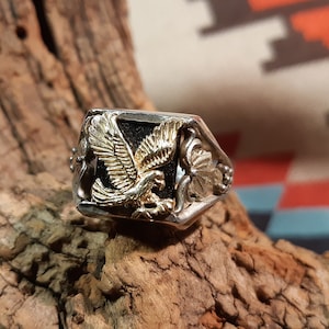 Op de afbeelding: Een zilveren ring met een zwarte steen en een gouden adelaar ontwerp.