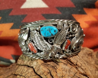 May include: A silver bracelet with a turquoise and coral inlay. The bracelet features a detailed design of a flying eagle.