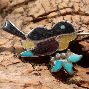 May include: A silver pin featuring a black and yellow bird with turquoise accents. The bird is perched on a branch with a turquoise butterfly.