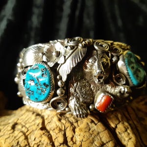 Può includere: Un bracciale a polsino in argento con accenti turchesi e corallo. Il bracciale presenta un design dettagliato di orso al centro, circondato da motivi floreali e a foglia. Le pietre turchesi sono di forma ovale e incastonate in argento.
