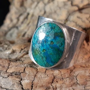 Könnte beinhalten: Ein Silberring mit einem großen, oval geformten Stein. Der Stein weist eine lebendige Mischung aus Blau- und Grüntönen auf. Der Ring hat ein breites Band, das sich um den Finger legt. Der Ring ist vor einem strukturierten Holzgrund platziert.