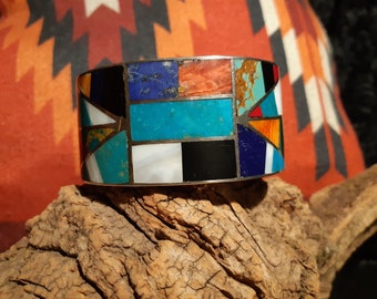 May include: A silver cuff bracelet with a geometric design made of turquoise, lapis lazuli, coral, and other stones.