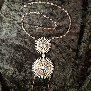 Könnte beinhalten: Eine silberfarbene Halskette mit zwei kreisförmigen Anhängern. Jeder Anhänger hat einen zentralen Türkisstein, der von strahlenförmigen Silberelementen umgeben ist. Die Halskette hat eine filigrane Kettengliederung, vor einem dunklen, strukturierten Stoffhintergrund.