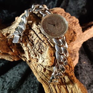 Puede incluir: Un reloj de pulsera plateado con esfera de moneda y una gruesa correa de eslabones. La esfera del reloj presenta un diseño en relieve. El reloj se exhibe sobre un trozo de madera desgastada, con un fondo oscuro.