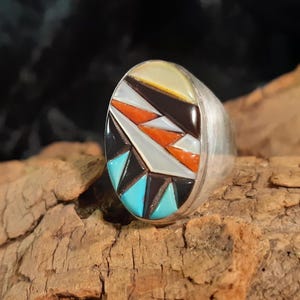 Könnte beinhalten: Ein silberner Ring mit einem bunten Einlegewerk aus Türkis, Rot, Weiß und schwarzen Steinen. Die Steine sind in einem geometrischen Muster angeordnet.
