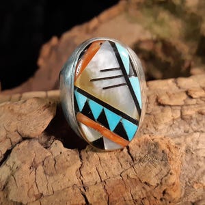 Könnte beinhalten: Ein silberner Ring mit einem Einlegemuster aus Türkis, Schwarz, Weiß und Orange. Der Ring hat ein geometrisches Muster mit einem zentralen weißen Stein.
