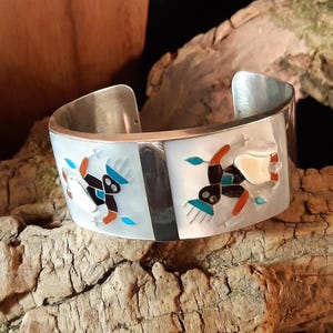 Op de afbeelding: Een brede zilveren manchetarmband met ingelegde ontwerpen. De armband heeft een witte achtergrond met kleurrijke ingelegde figuren, waaronder zwarte, bruine, rode en turquoise elementen. Een zwarte streep verdeelt de twee zijden.